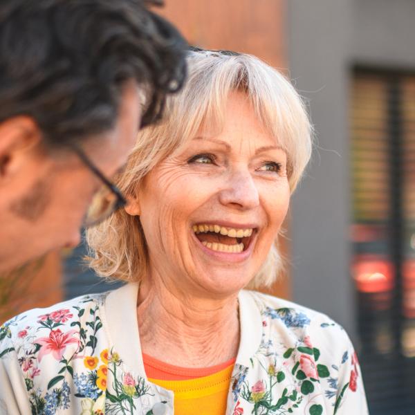 Une femme agée arborant un grand sourire au pied d'un immeuble